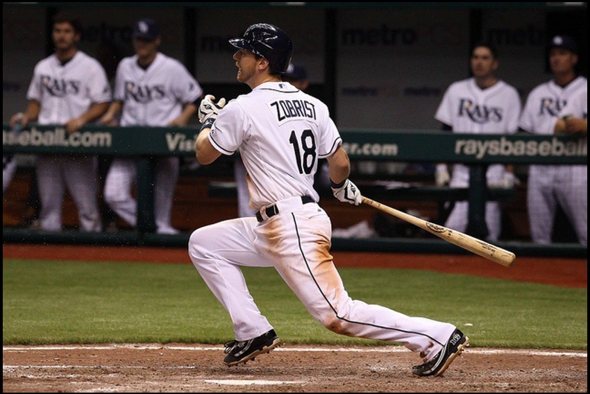 A baseball batter wearing jersey number 18, with players standing in the dugout behind him.